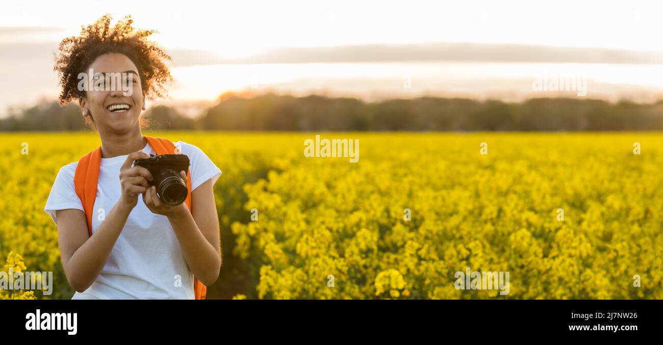 Panorama of beautiful biracial African American girl teenager female young woman smiling taking photographs with a camera at sunset or sunrise in a fi Stock Photo