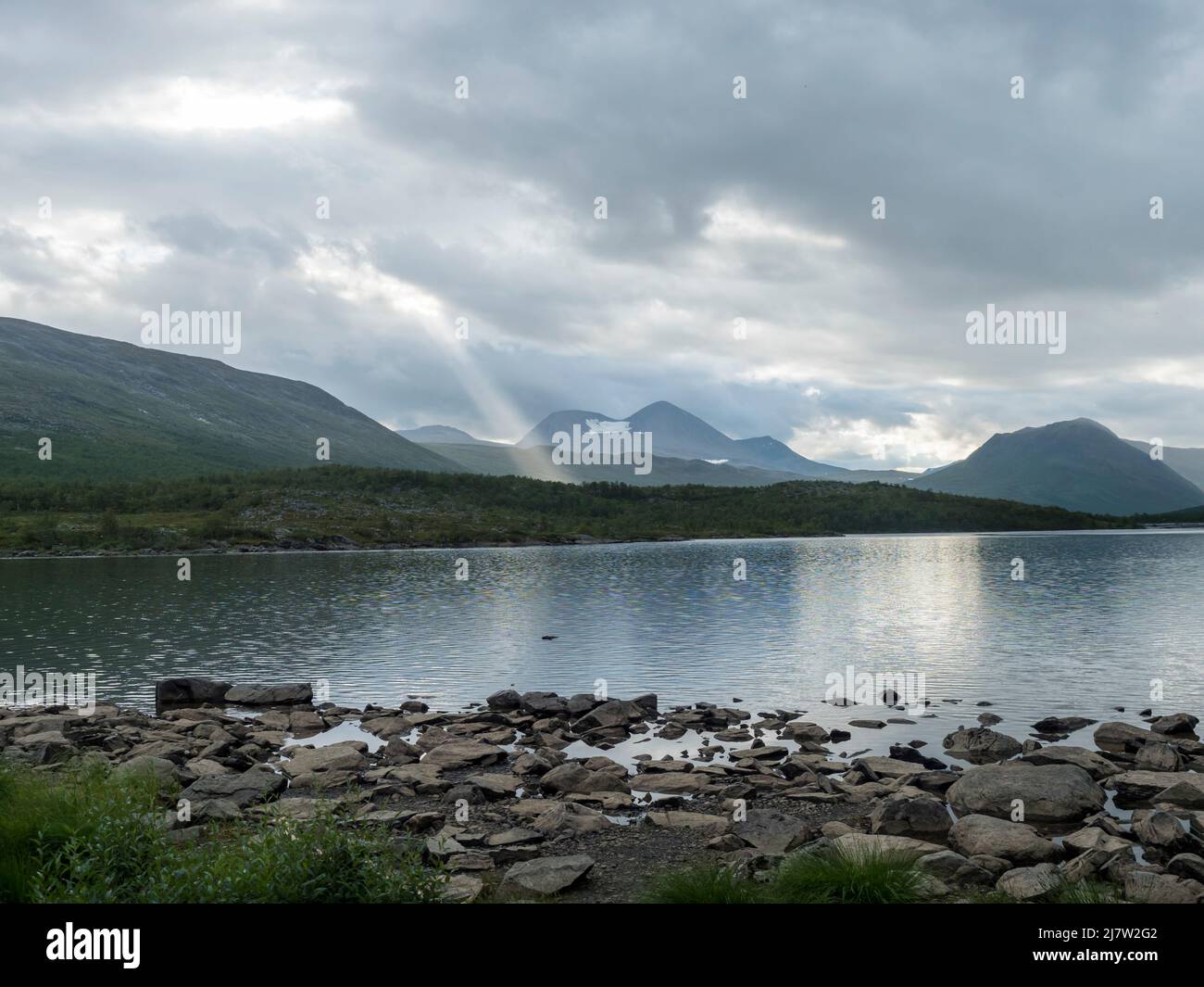 rocky shore of Tarra river. Tarraatno with green hills, birch tree forest and mountians at Padjelantaleden hiking trail. Summer cloudy evening at Stock Photo
