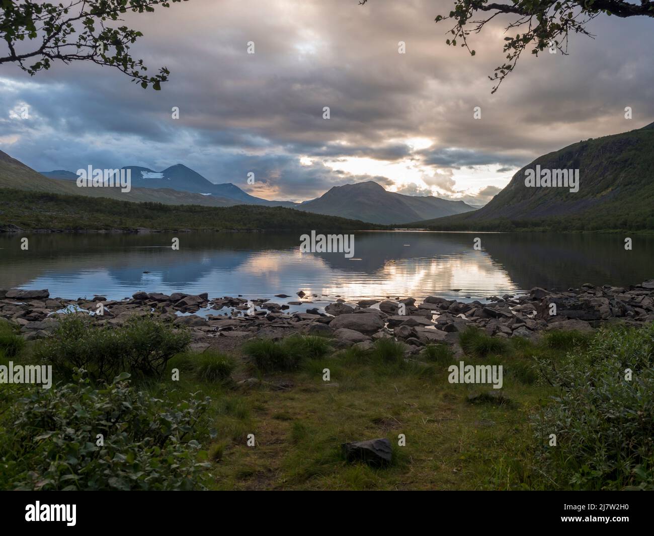 Sunset at rocky grassy shore of Tarra river. Tarraatno with green hills, birch forest and mountians at Padjelantaleden hiking trail. Summer cloudy Stock Photo