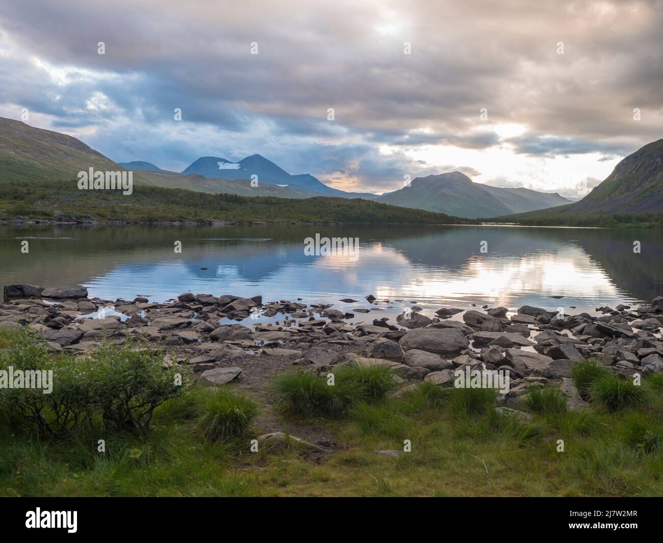 Sunset at rocky grassy shore of Tarra river. Tarraatno with green hills, birch forest and mountians at Padjelantaleden hiking trail. Summer cloudy Stock Photo