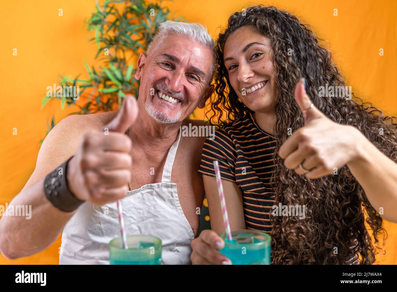 father and daughter drinking a blue cocktail doing the thumb up sign with their hands - studio shot on yellow background Stock Photo