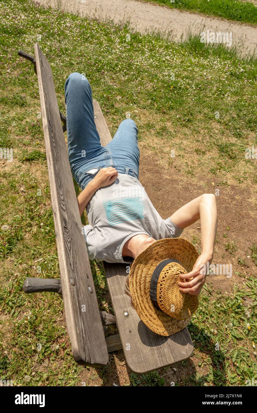 Woman with brown hair, gray t-shirt, jeans and straw hat lying down on a wooden park bench in the nature, relaxing, taking a nap, sunbathing, high ang Stock Photo