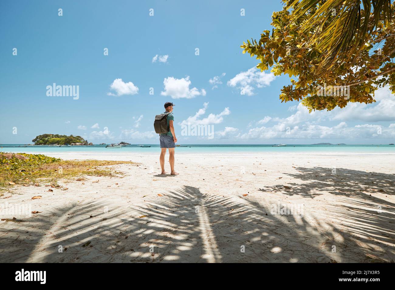 Man with backpack walking on white sand beach against idyllic seascape. Praslin island, Seychelles. Stock Photo