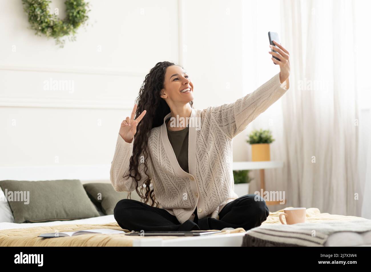 Young smiling woman sitting on bed taking selfie Stock Photo