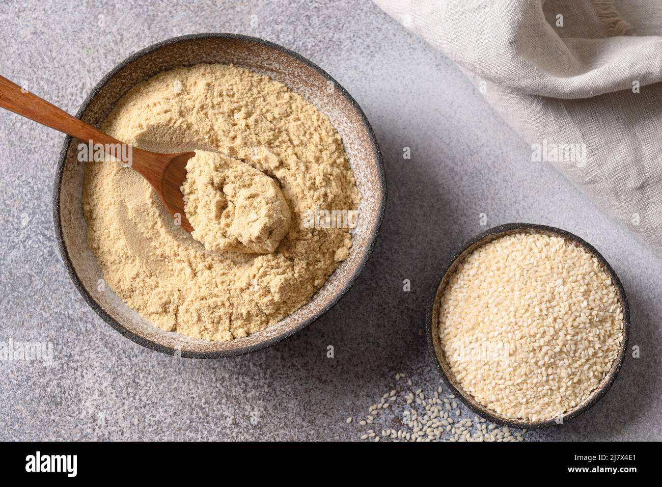 Sesame flour in bowl and white sesame seeds on gray background for cooking gluten-free and low carbohydrate dessert. Sesame flour includes ample miner Stock Photo