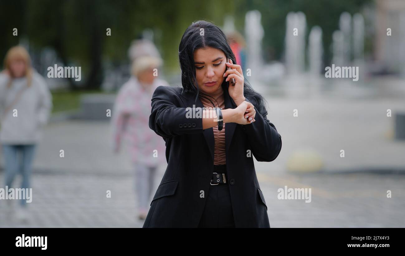 Young businesswoman excited calls on mobile phone angry that person late for meeting at scheduled time looking at wrist watch with irritation hispanic Stock Photo