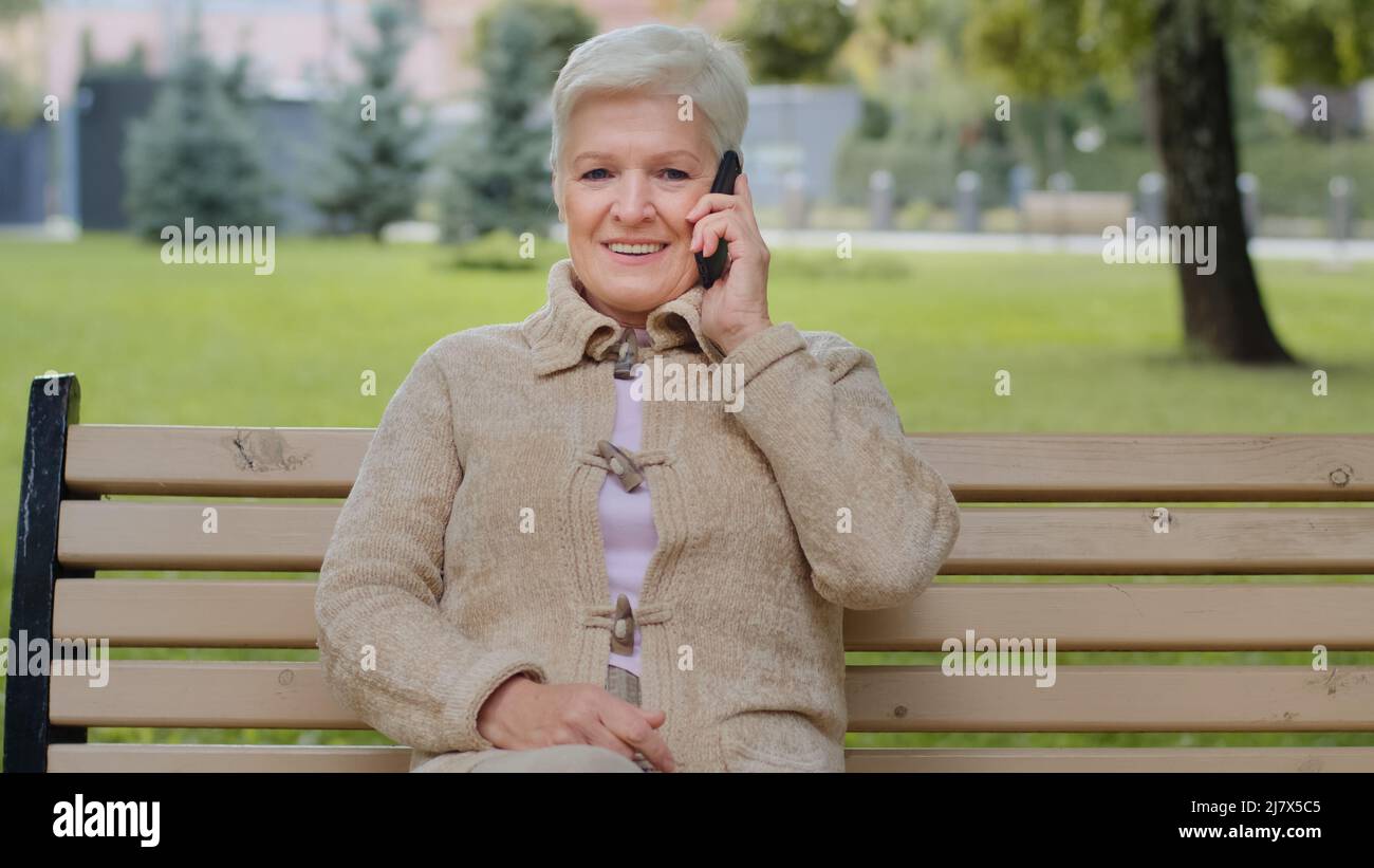 Smiling elderly gray-haired lady with beautiful face sitting on park bench takes phone call, woman of retirement age holds smartphone in hand answers Stock Photo
