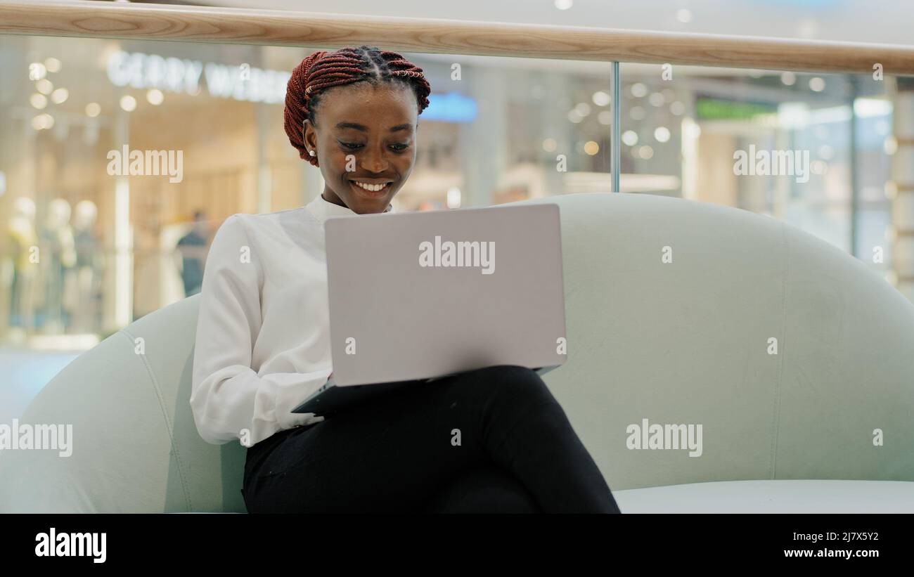 Happy smiling girl african american student female business woman freelancer sitting on sofa in office shopping center chatting typing on laptop Stock Photo