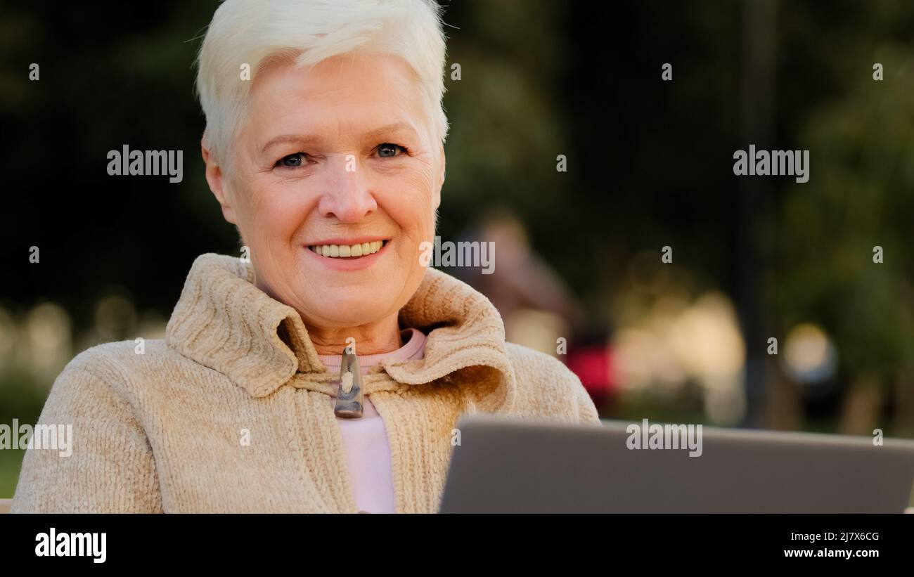 Smiling senior female of retirement age working at laptop reading news online, elderly lady texting message at pc outdoor. Happy aged woman using a Stock Photo
