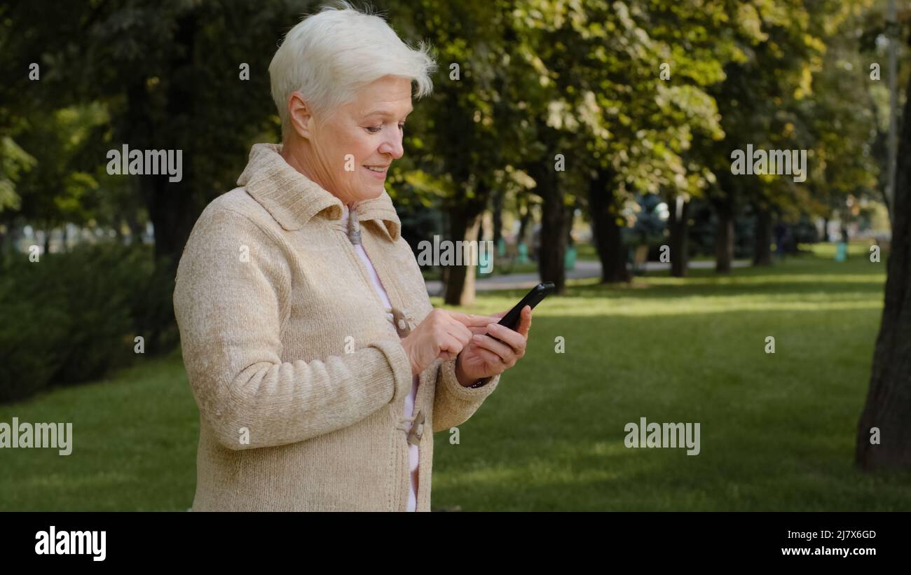 Closeup gray-haired Mature woman in casual clothes walking outdoors in city park uses cellphone modern gadget smartphone, browses message Stock Photo