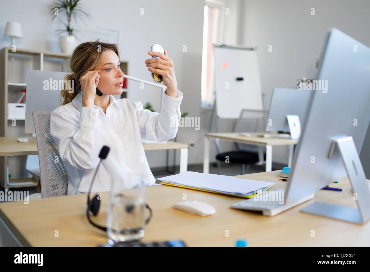 Elegant office woman sits at her workplace and looks in the mirror to touch up her makeup Stock Photo