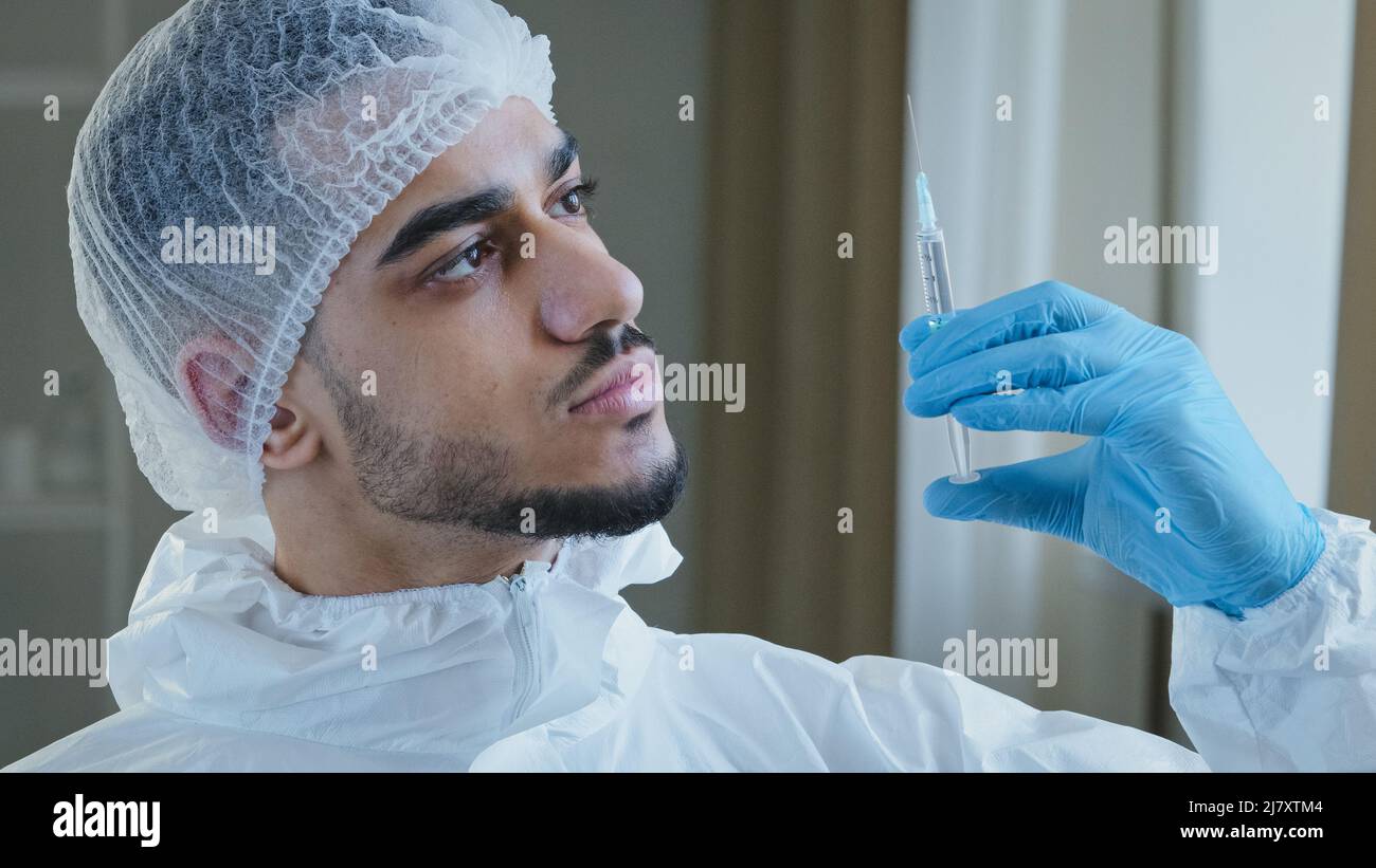 Young arabic male hispanic doctor practitioner surgeon in special medical protective uniform wears rubber gloves holds syringe with medicine vaccine Stock Photo