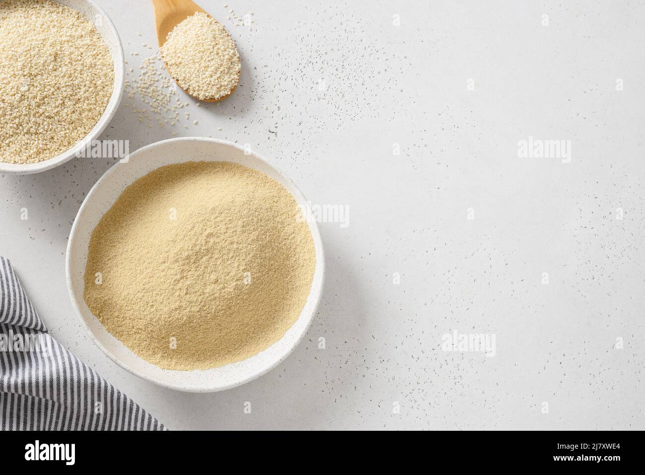 Sesame flour in bowl and white sesame seeds in wooden spoon on white background. View from above. Copy space. Good vegan source of protein, minerals, Stock Photo