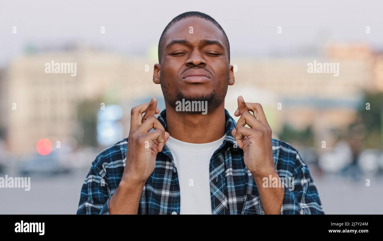 African American young hopeful man stands in city on street crossing fingers praying says prayer hopes for future possibility, hope faith concept Stock Photo