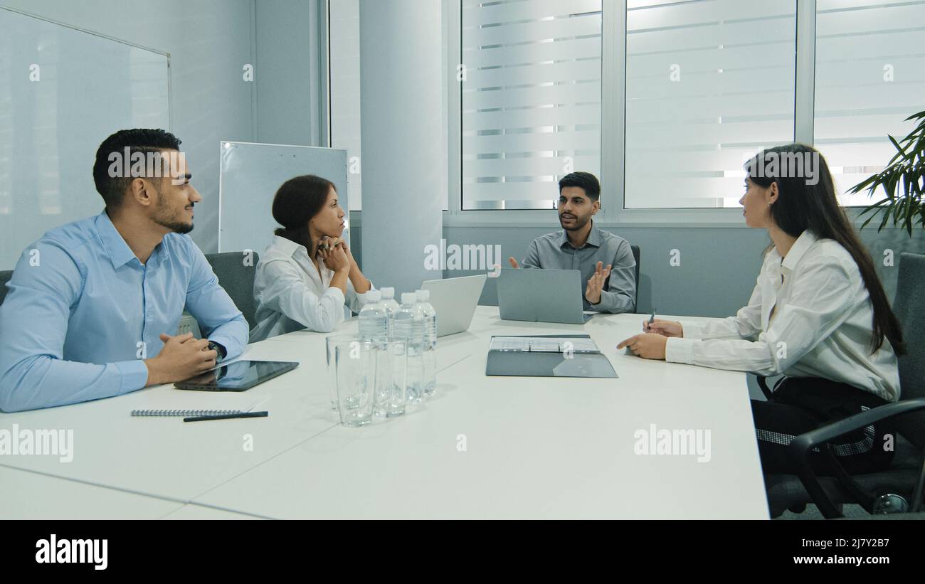 Young indian team leader sitting with colleagues, discussing issues at negotiations meeting. Skilled millennial diverse employees working together at Stock Photo