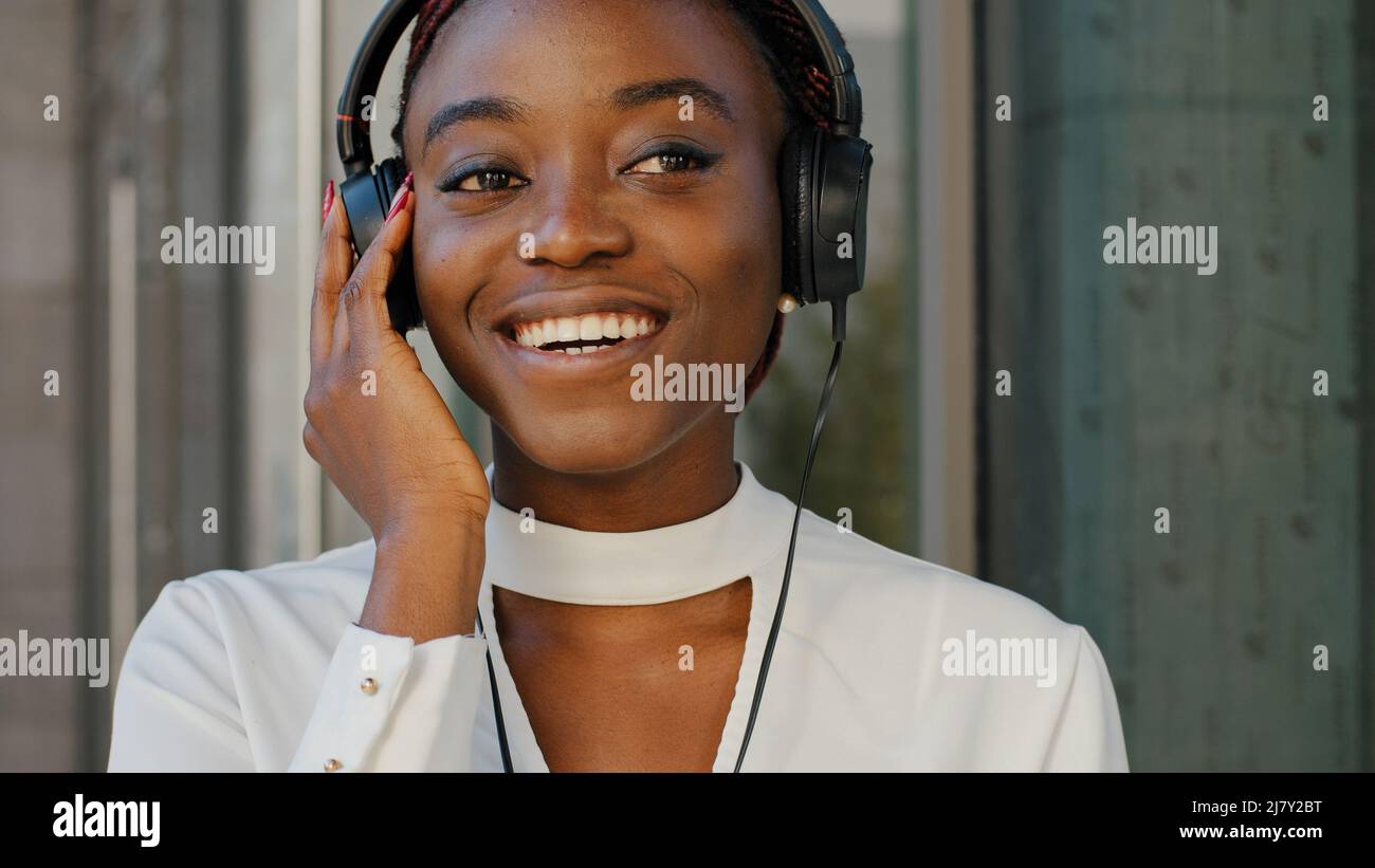 Portrait of young beautiful cheerful funny African American girl student tourist stands in headphones listening to music in city. Close-up woman Stock Photo