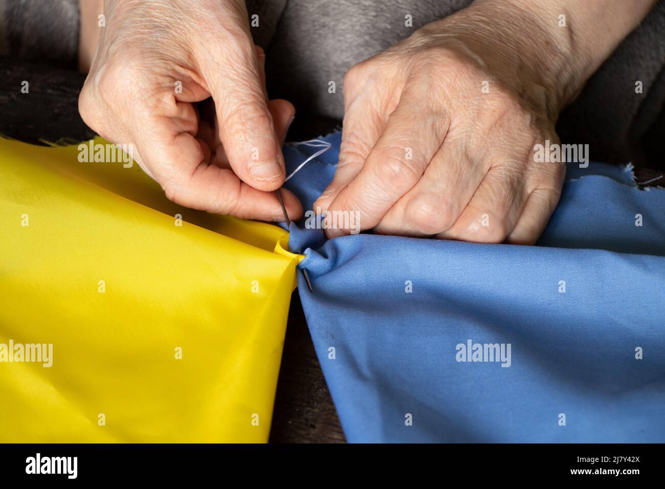 Female old Ukrainian woman hands sew the national flag of Ukraine yellow-blue at the table at home in Ukraine, patriot, stop the war in Ukraine 2022 Stock Photo