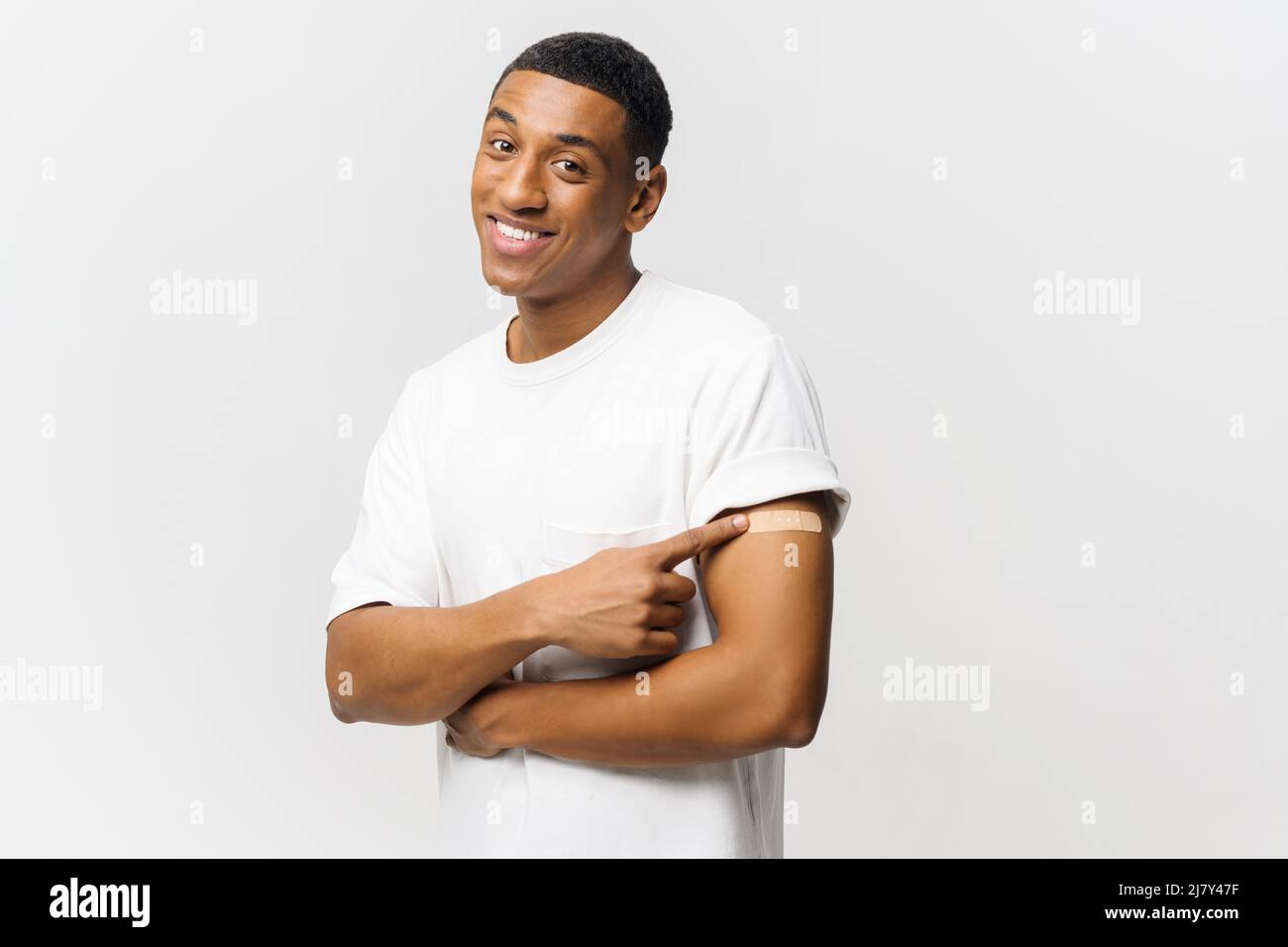 Smiling young African-American guy showing arm with band-aid after vaccine injection isolated on blue background, african-american man point finger at patch on his forearm Stock Photo