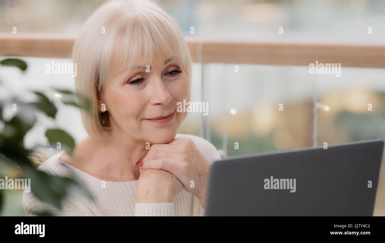 Happy calm mature middle aged woman sitting resting relaxing holding hands under chin looking at laptop screen browsing photos remembering past dreams Stock Photo