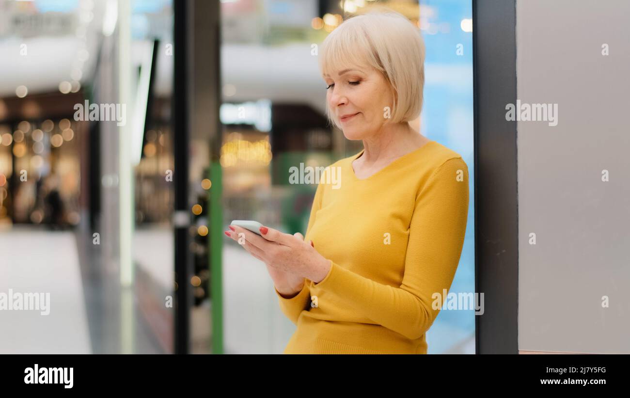 Smiling middle aged mature caucasian woman holding phone looking at smartphone screen sending message checking email using mobile application browsing Stock Photo