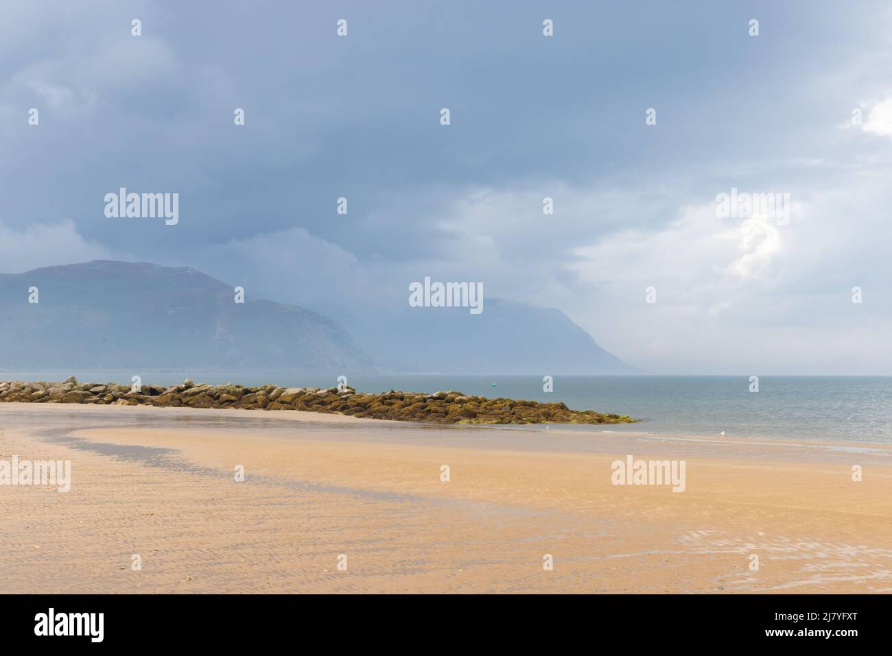 The sands and breakwater on Llandudno's West Shore looking towards Conwy Sands and Bay. In the distance dark clouds over Penmaen-bach threaten rain. Stock Photo