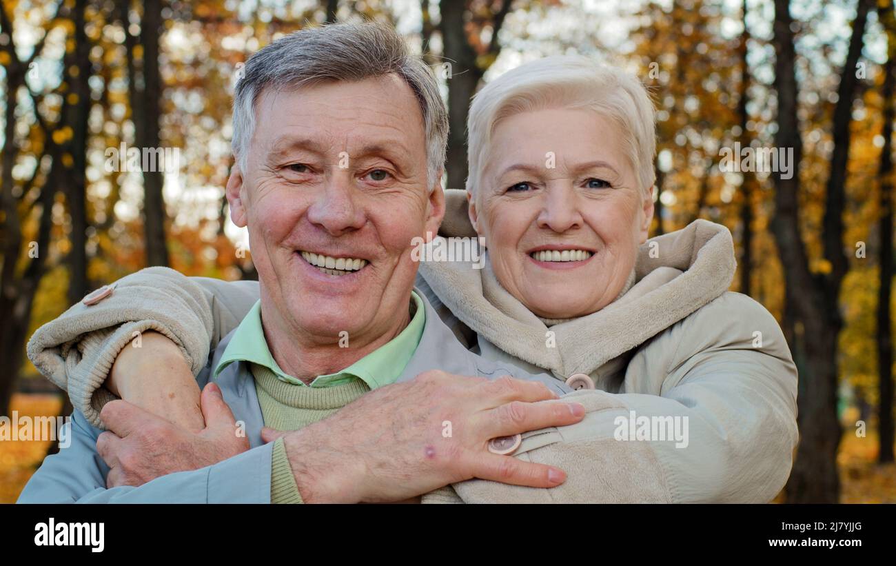 Adult old woman hug beloved husband by shoulders elderly married couple hugging in autumn park grandparents smiling looking at camera then each other Stock Photo