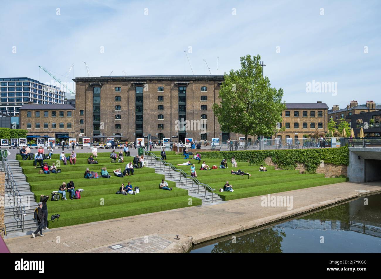 People relaxing on the grass terrace seating area on the bank of the Regents Canal on a sunny springtime day at Coal Drops Yard. London, England, UK. Stock Photo