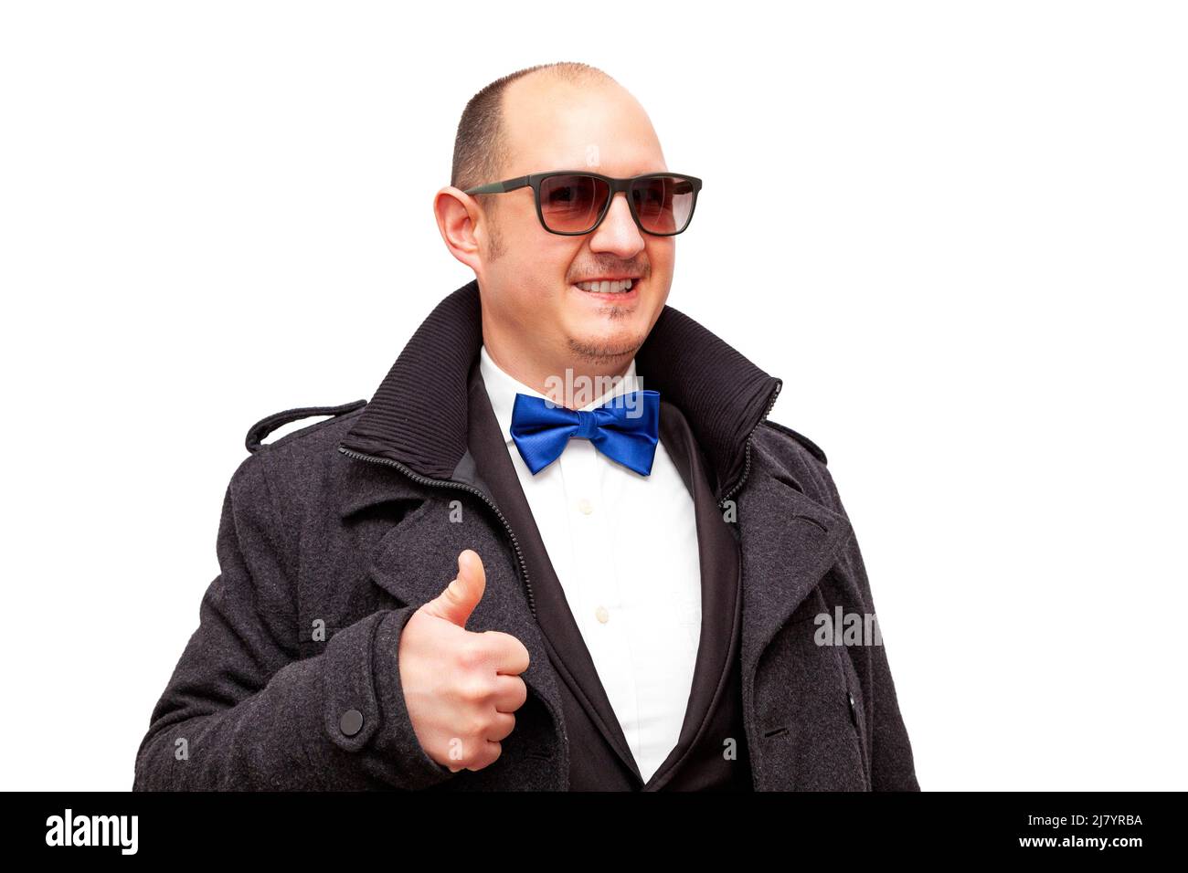 A bald adult man wearing sunglasses and dressed in smart clothes and a blue bow tie is holding up one of his thumbs while smiling. The male is on a wh Stock Photo