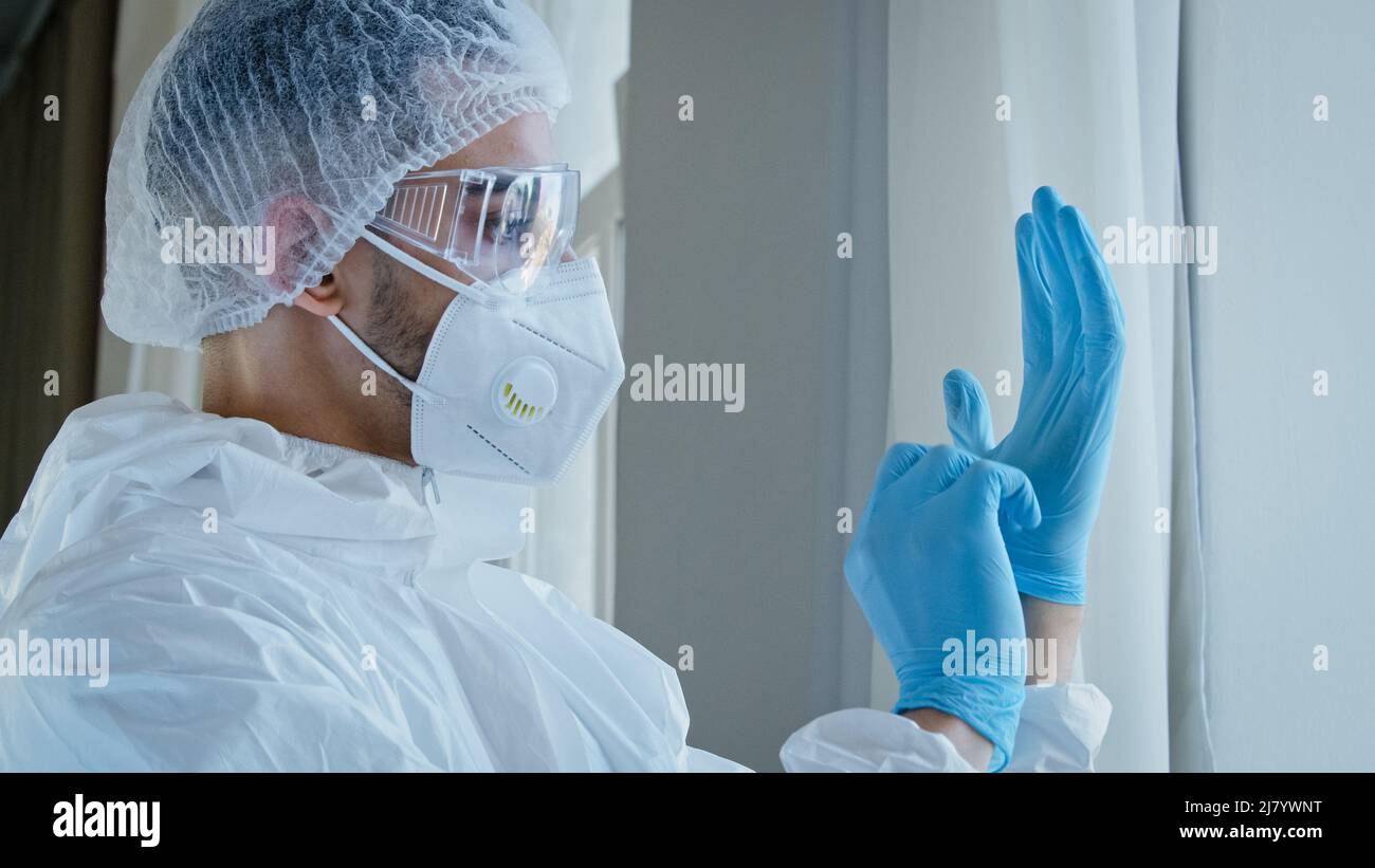 Young man doctor male nurse medical worker in special protective suit uniform with glasses and cap puts on rubber blue gloves on hands preparing Stock Photo