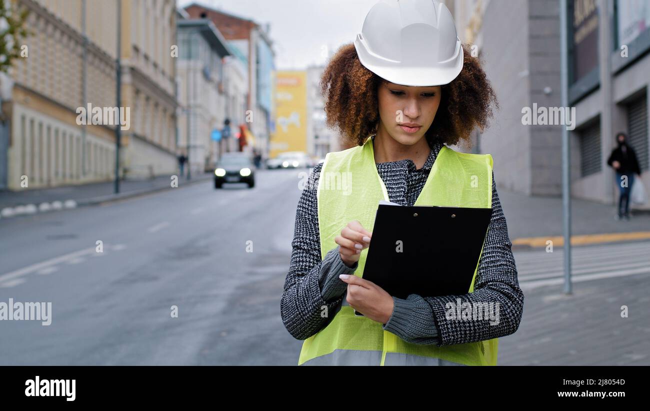 Portrait young african american beautiful female engineer wearing safety vest and hardhat standing in city near road traffic writing data Stock Photo