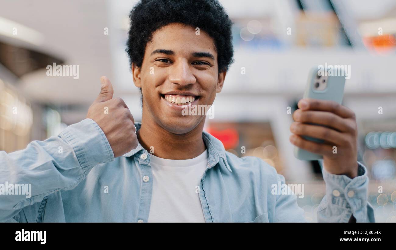 Portrait satisfied happy african guy man with phone smartphone modern gadget looking at camera shows like thumbs up recommends wifi cellular Stock Photo