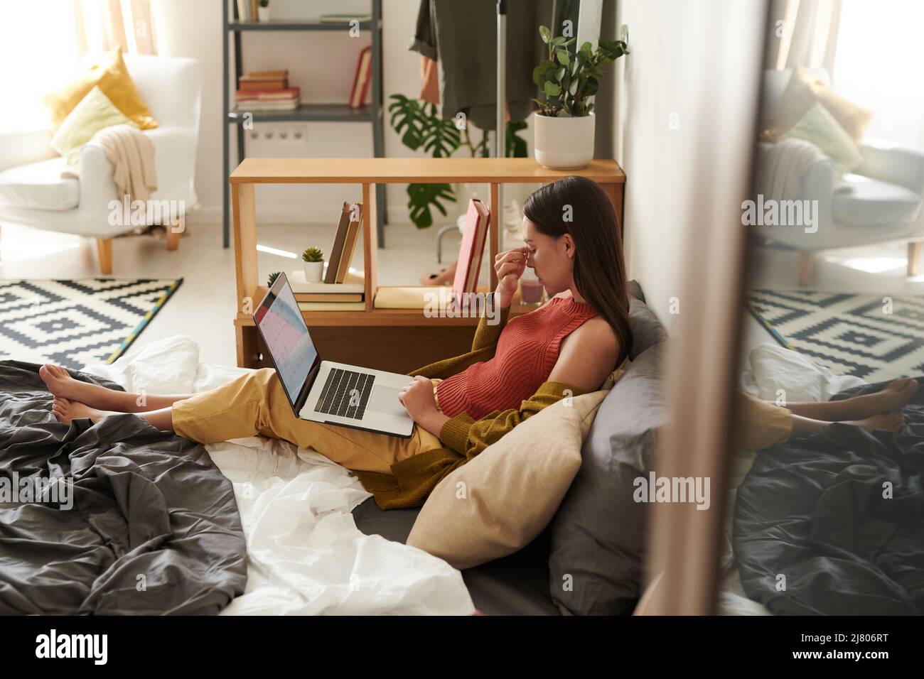 Overworked young woman with brown hair sitting with laptop on bed and