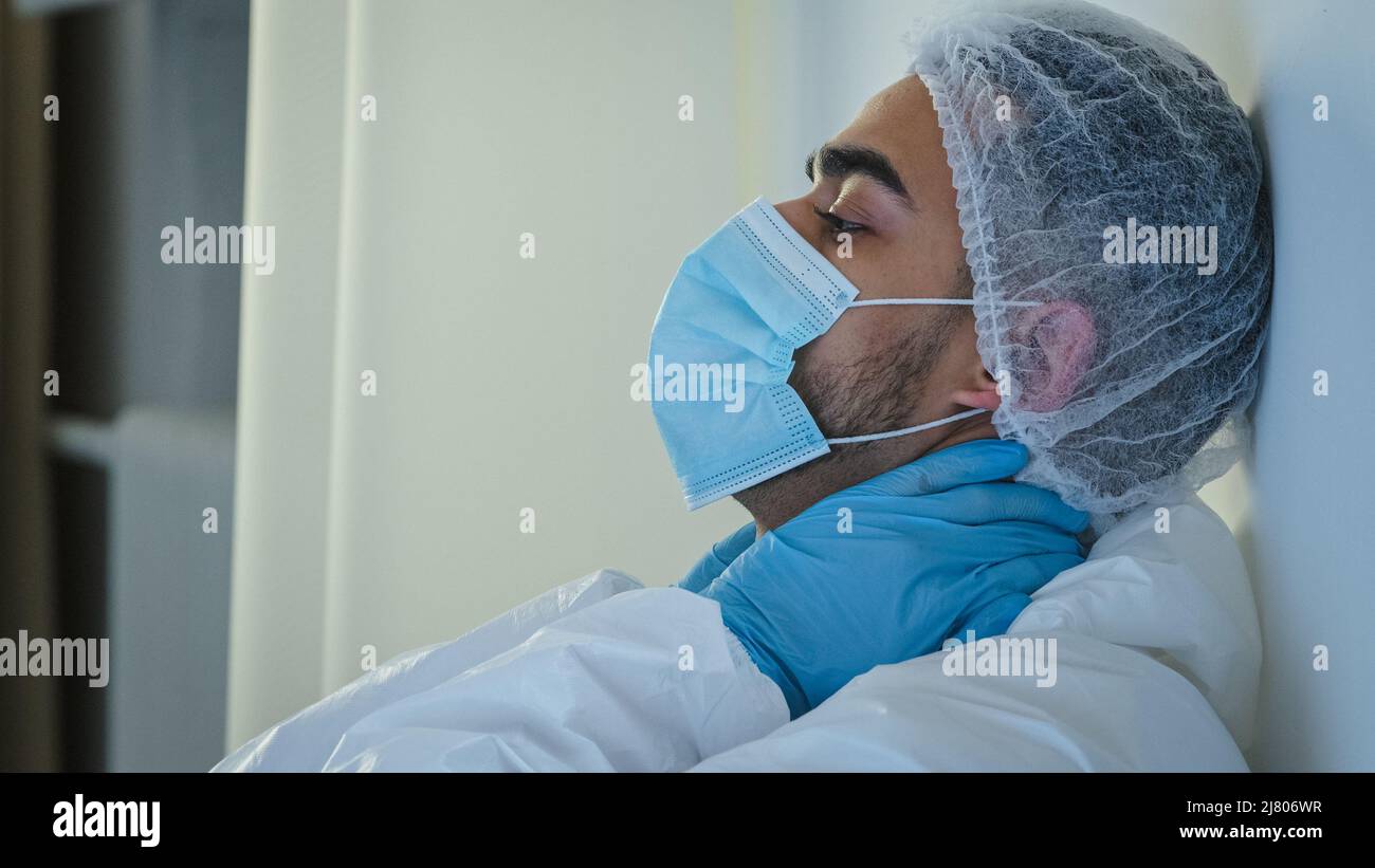 Side view male doctor medical worker surgeon in protective mask sitting on floor in clinic near wall holding neck with hands feeling tired exhaustion Stock Photo