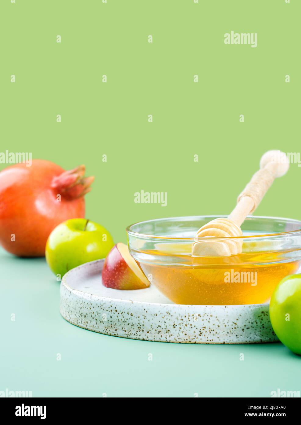 glass saucer with fresh honey, honey spoon, apple and pomegranate on a bright green background. Concept Jewish New Year Happy holiday Rosh Hashanah Stock Photo