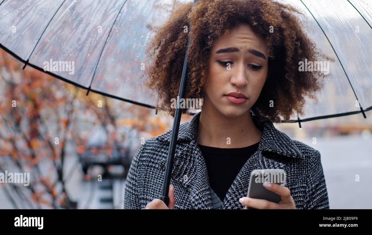 African American woman curly girl with transparent umbrella stands on autumn street in rain looks at mobile phone upset sadness worrying about low Stock Photo