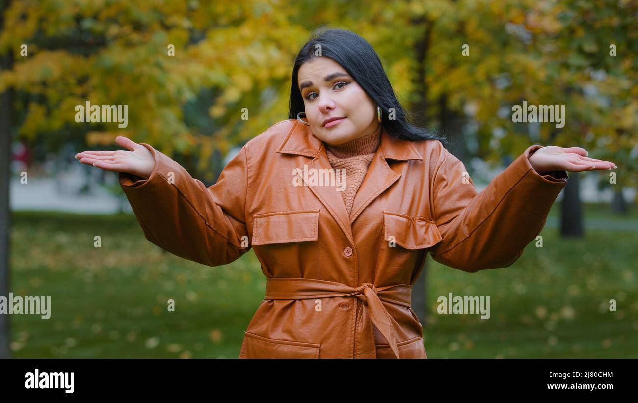 Young puzzled upset hispanic woman standing outdoors in park shrugging shoulders doubtful looking at camera unsure does not know what to answer Stock Photo