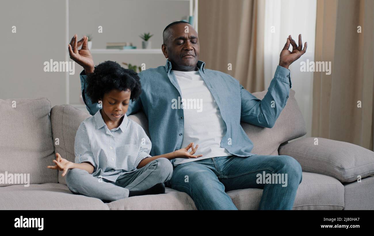 African american father and child sitting on sofa with closed eyes mature dad teaches little daughter to meditate manage emotion calm down do yoga Stock Photo