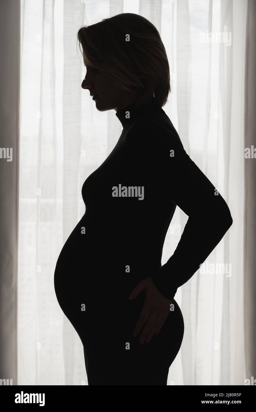 Side view of silhouette pregnant young woman standing against window at home. Portrait expecting woman in darkroom. Stock Photo