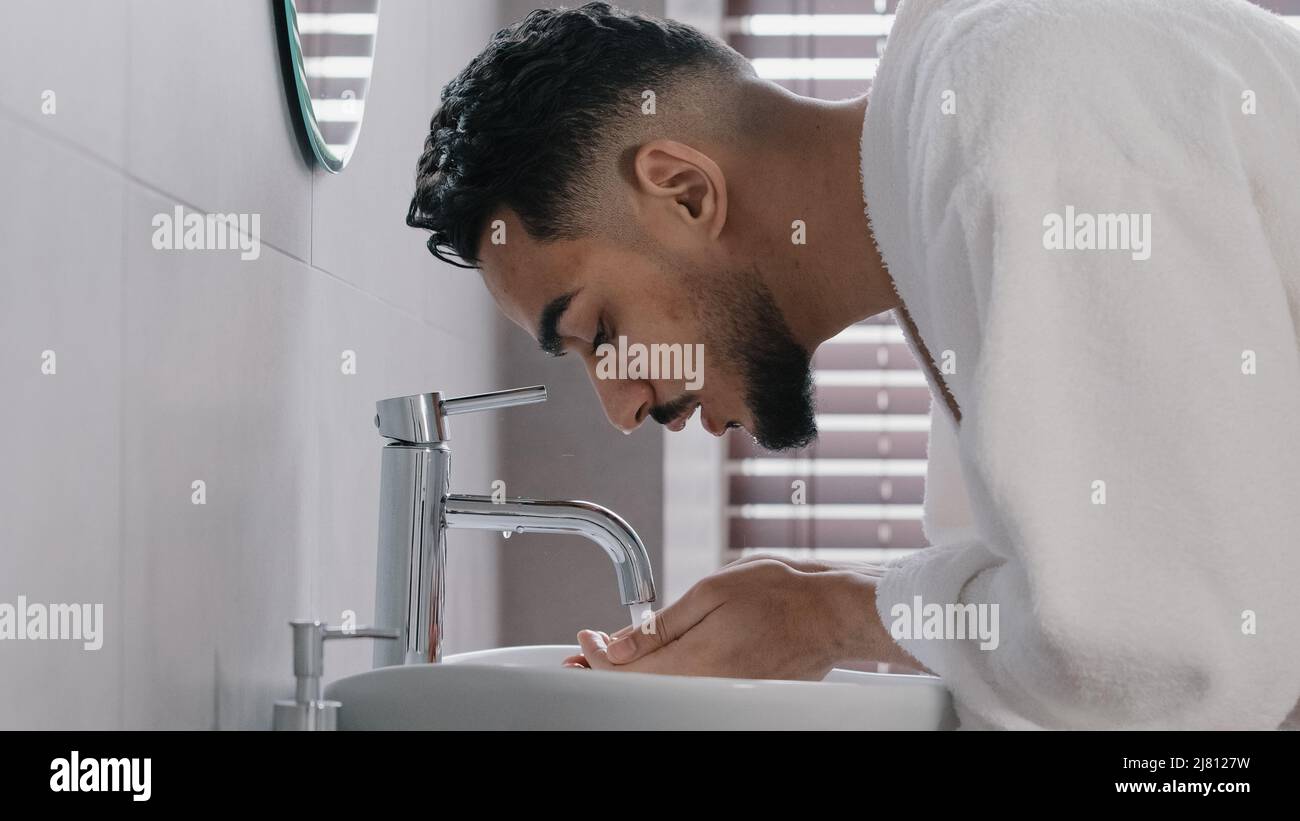 Side view arab spaniard indian arab man guy opens faucet near washbasin at home in hotel bathroom washes face with hot cold water morning beauty Stock Photo
