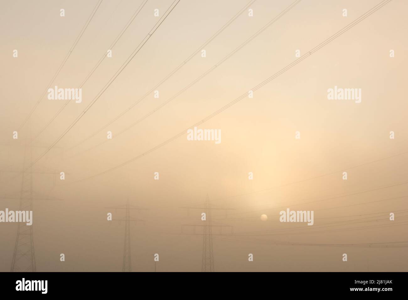 Setting sun in the mist behind three power poles with the foggy orange sky as background Stock Photo