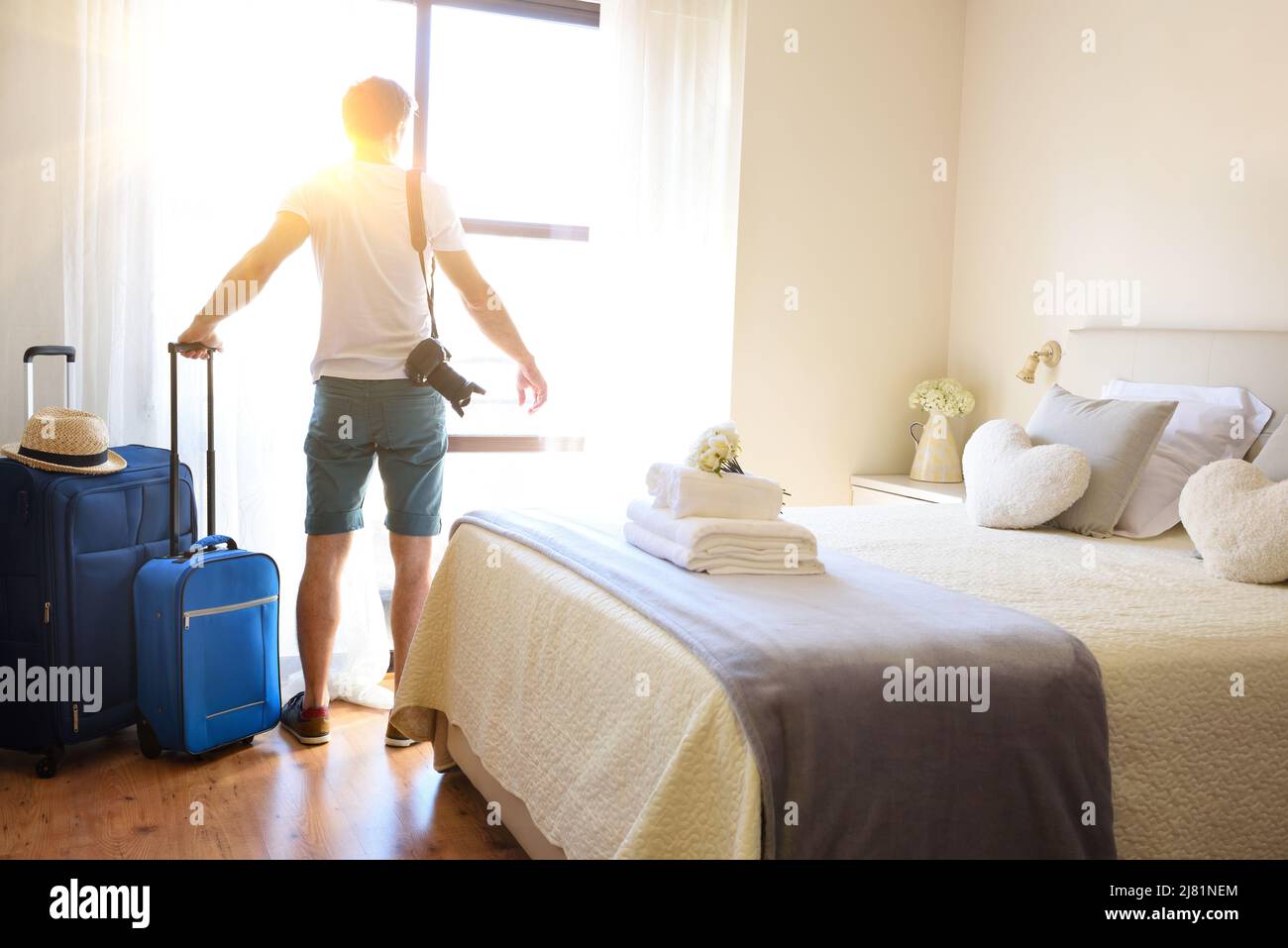 Man on vacation with suitcases on his backs looking out a window in a bright hotel room. Stock Photo