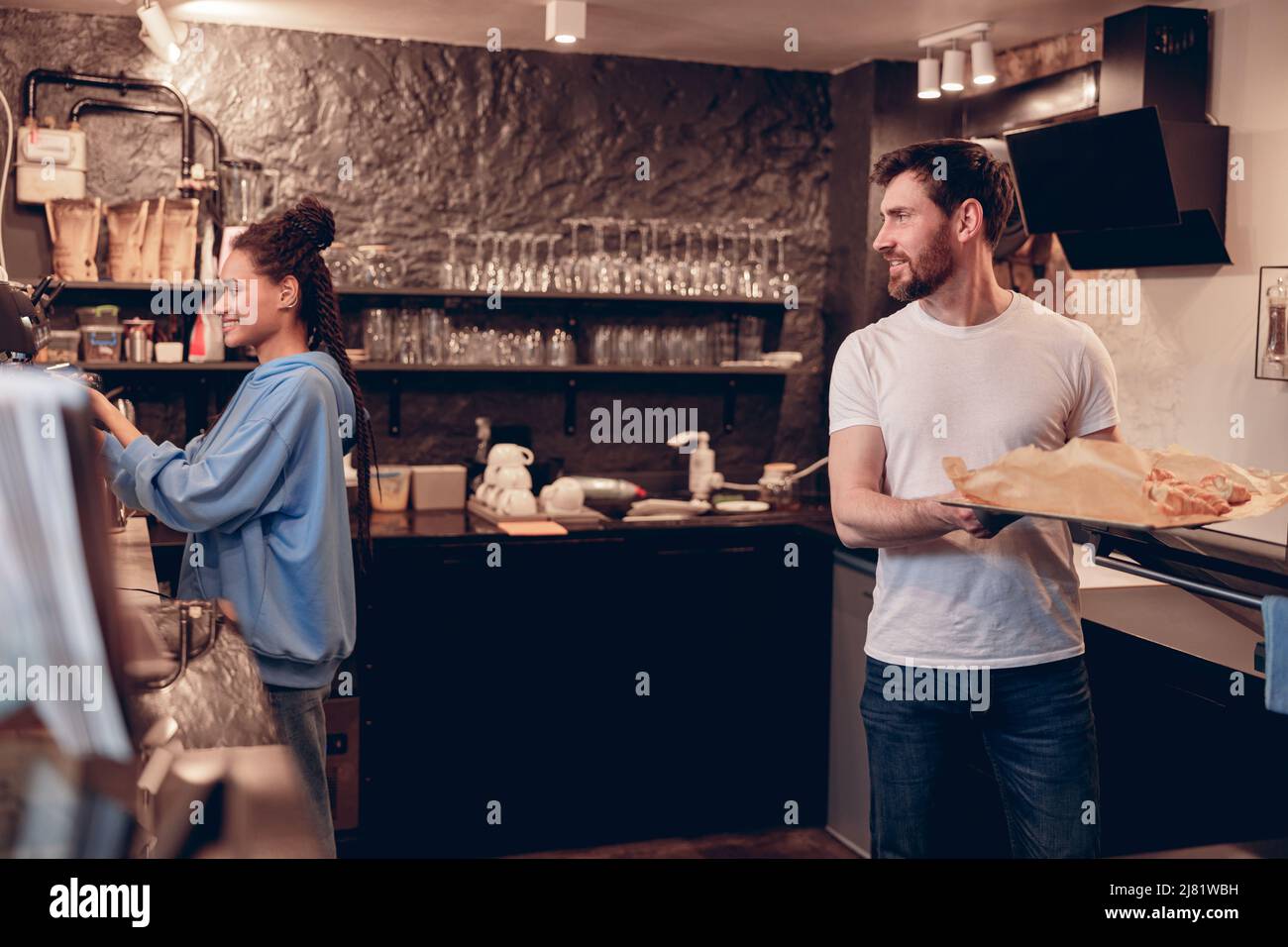 Young cafeteria workers working at bar kitchen. Female making coffee, male baking croissants. Stock Photo