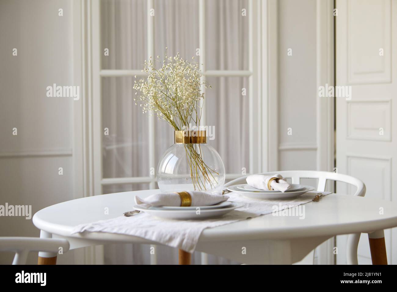 Elegant glass vase with gentle babys breath flowers and ceramic plates with rolled napkins served on round table in white dining room Stock Photo