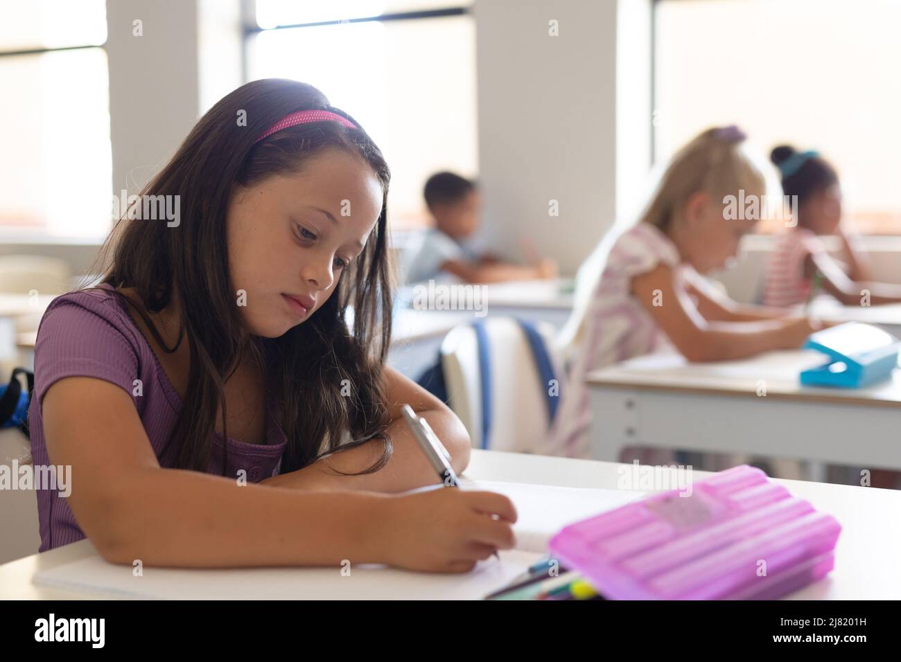 Caucasian elementary schoolgirl writing on book while studying at desk ...