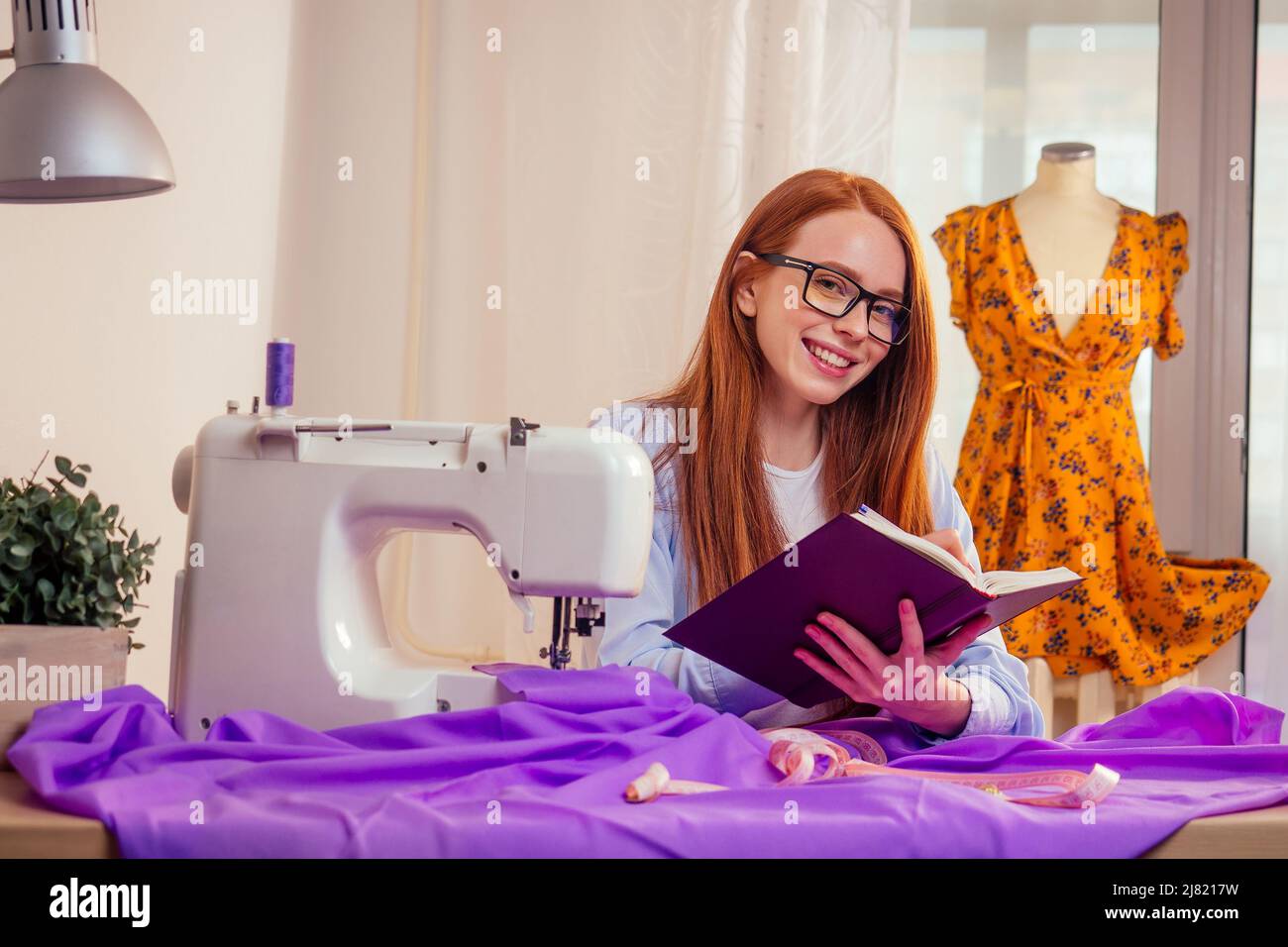 creative designer red haired woman wearing glasses, planning new collection clothes and writing notes in office workroom Stock Photo