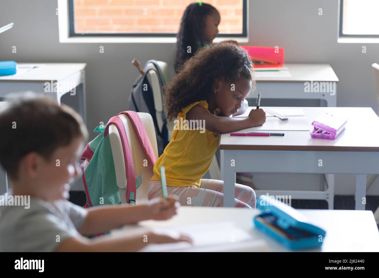 Multiracial elementary school students writing on book while studying at desk in classroom Stock Photo