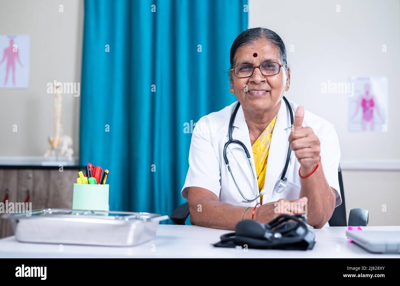 Happy senior woman doctor showing thumbs up by looking at camera at hospital with copy space - concept of successful, professional occupation and Stock Photo