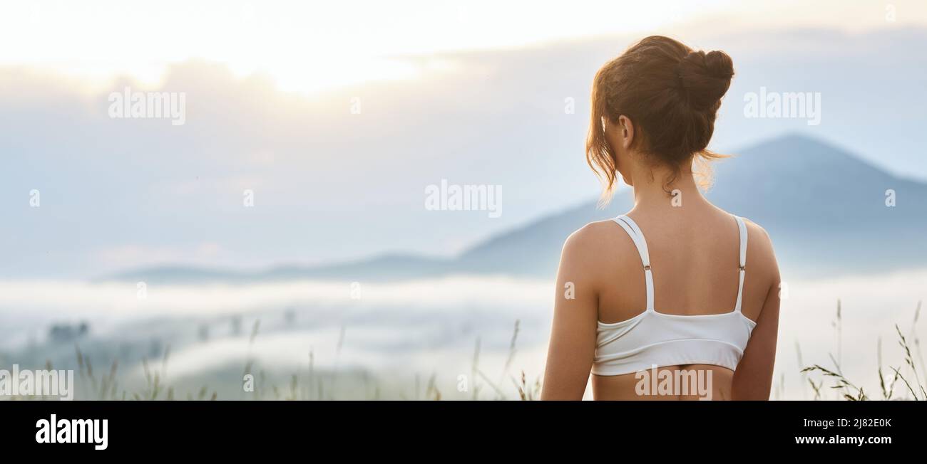 Back panoramic view of woman looking forward in hills. Female with hair bun, wearing white top, enjoying beautiful landscape, dreaming. Concept of harmony with nature. Stock Photo