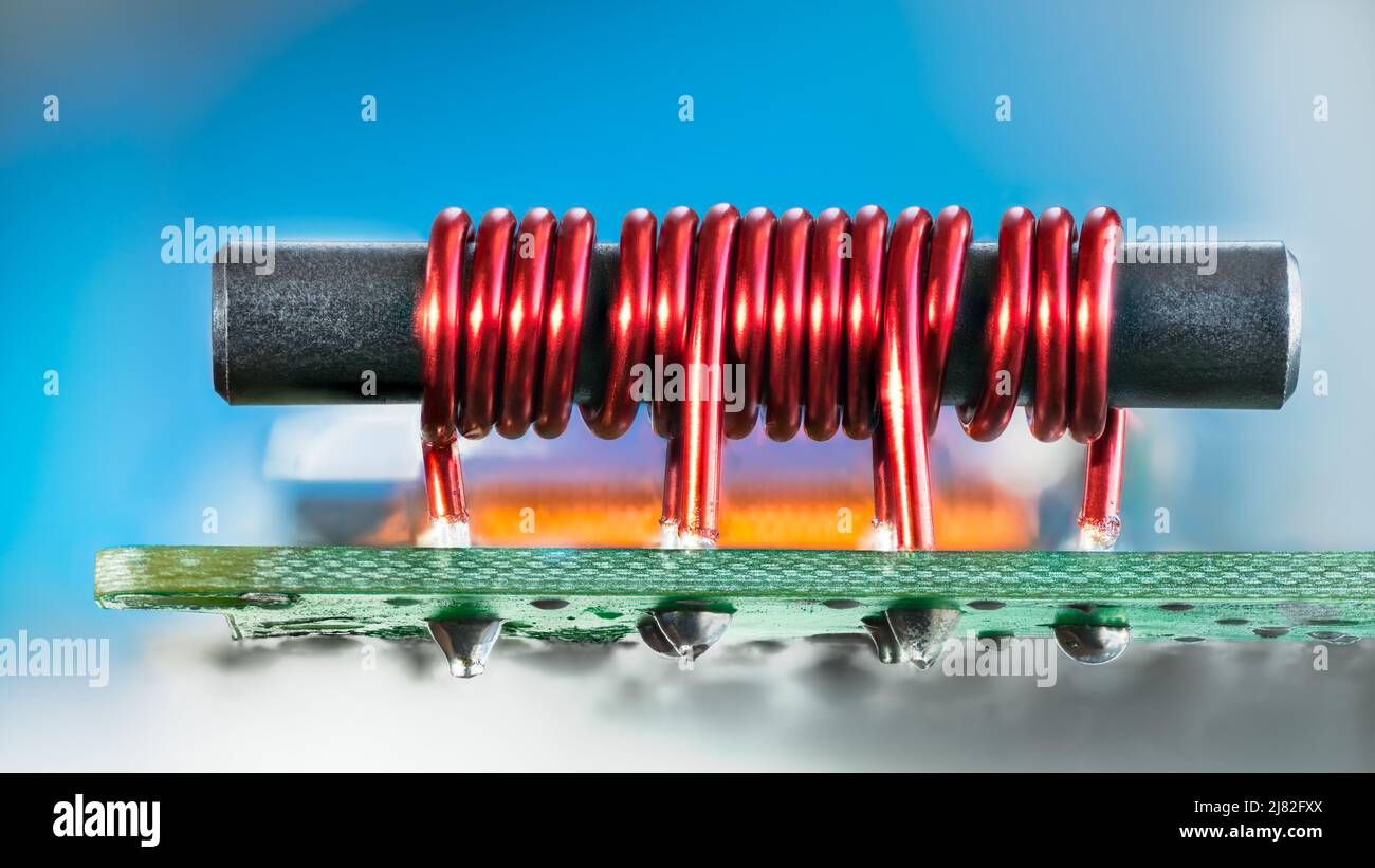 Electronic transformer with ferromagnetic core and coil copper wire on PCB. Closeup of red cylindric inductor soldered on green circuit board. TV amp. Stock Photo
