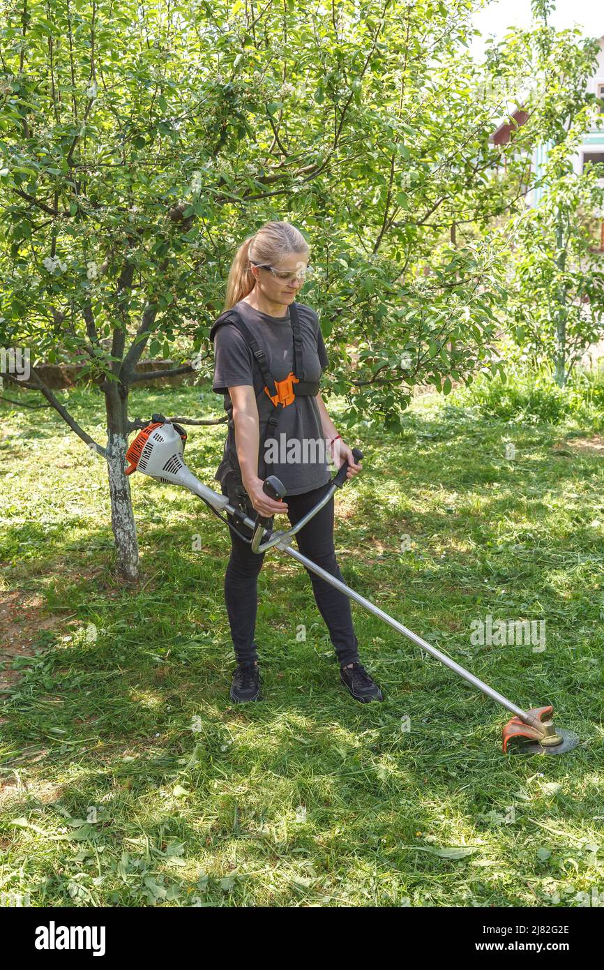 A woman mows the grass in the garden with a gasoline trimmer. She wore